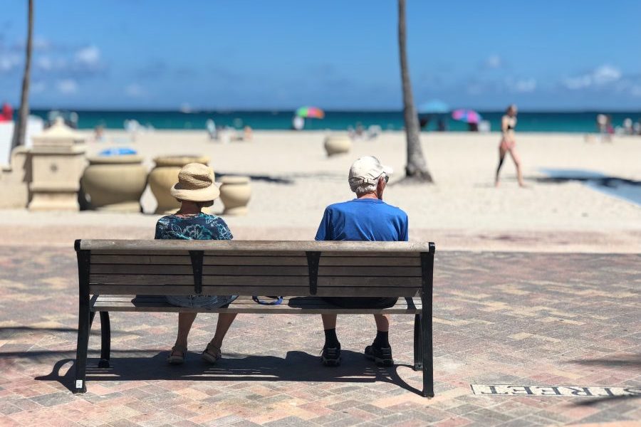Back view of two seniors sitting on a bench, with the beach in the background.