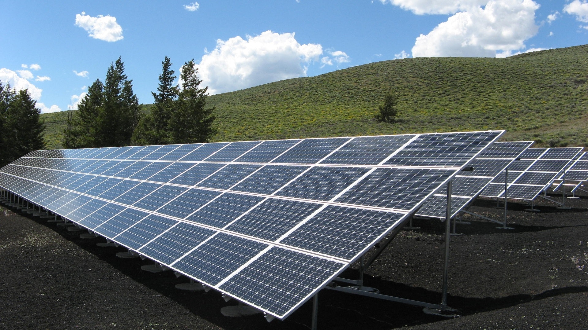 Solar panels in a green field with a few trees, clouds, and a hill in the background.